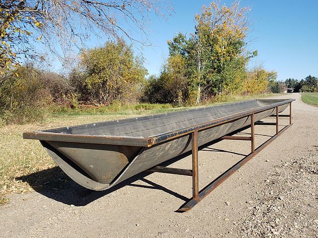 a long metal boat sitting on the side of a road next to a tree filled field