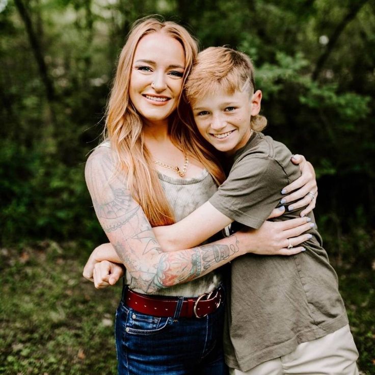 a woman hugging a boy with tattoos on his arms in the middle of a forest