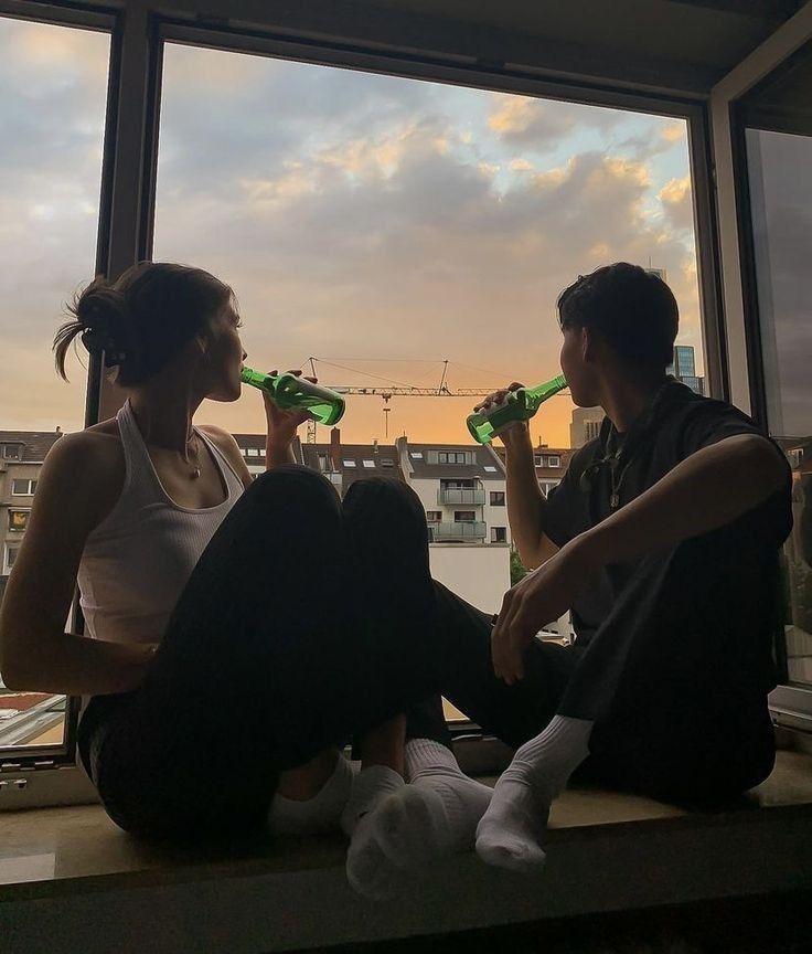 two people sitting on a window sill drinking from green bottles with buildings in the background