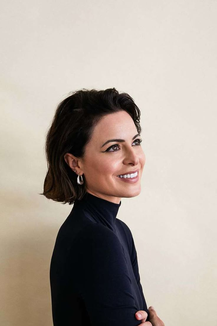 a smiling woman with her arms crossed, wearing a black top and silver earrings in front of a white wall