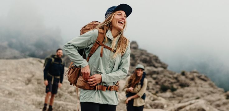 three people hiking up a mountain with backpacks on their back and one woman laughing