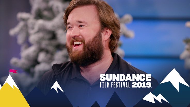 a man with long hair and beard smiling in front of snow covered mountains on the set of sundance film festival