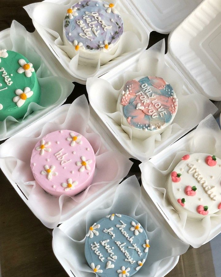 six decorated cookies in plastic trays on top of a wooden table with white and pink frosting