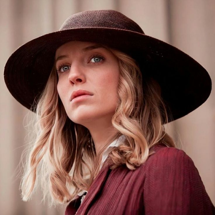 a woman with blonde hair wearing a brown hat and looking off into the distance while standing in front of a building