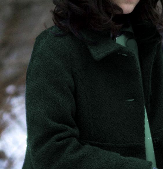 a woman wearing a green coat and holding a cell phone in her hand with snow on the ground behind her