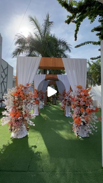 an outdoor ceremony setup with white drapes and orange flowers on the grass, surrounded by palm trees