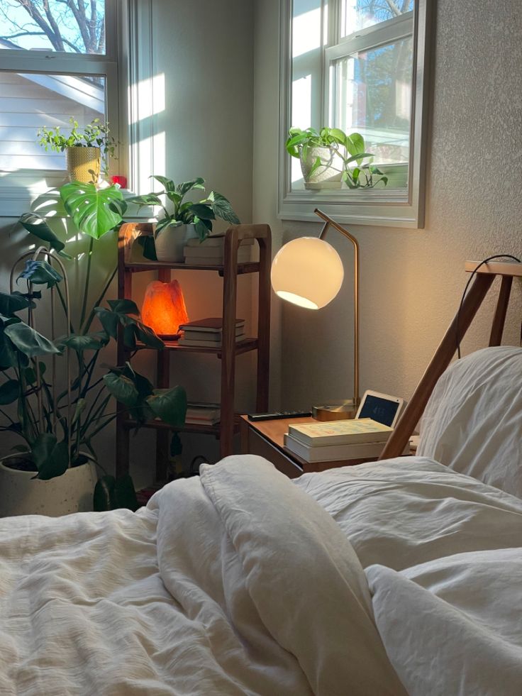 a bed with white sheets and pillows next to a window filled with potted plants