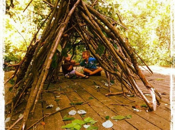 two people sitting in front of a wooden structure made out of sticks and branches with leaves on the floor