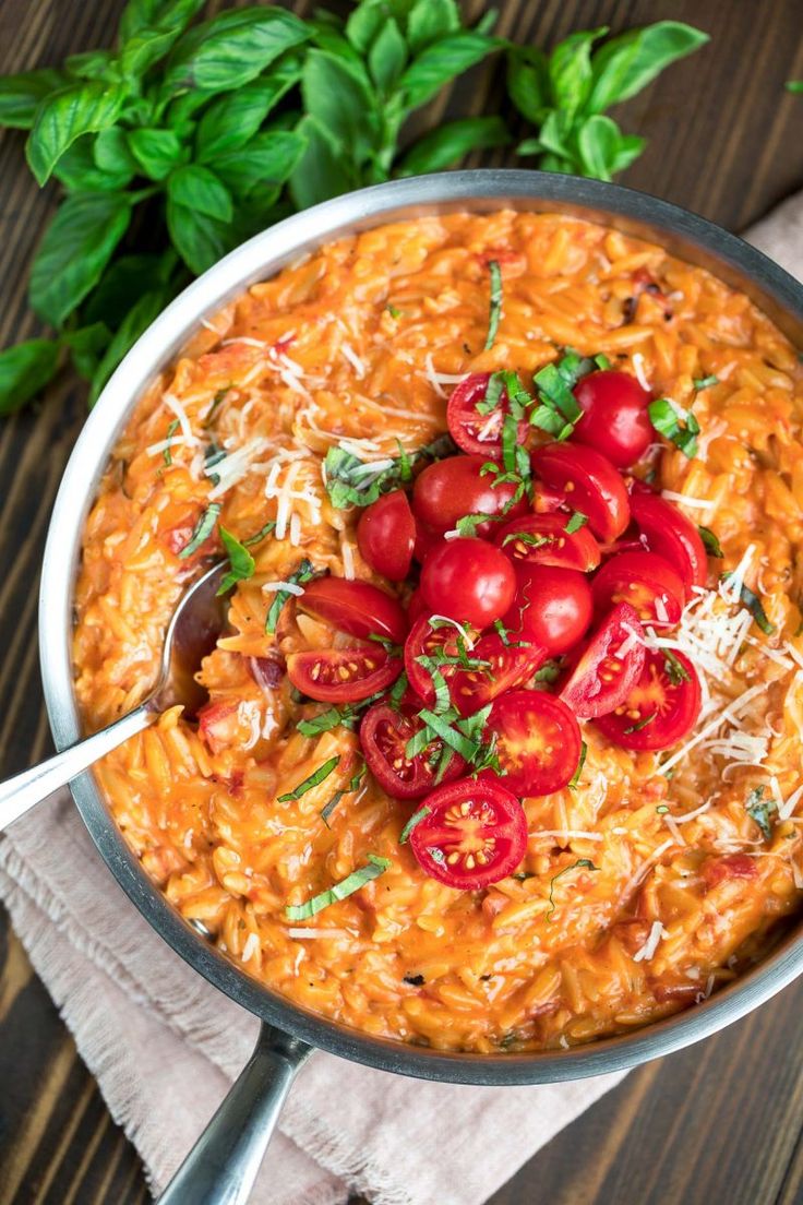 a pan filled with pasta and tomatoes on top of a wooden table next to basil leaves