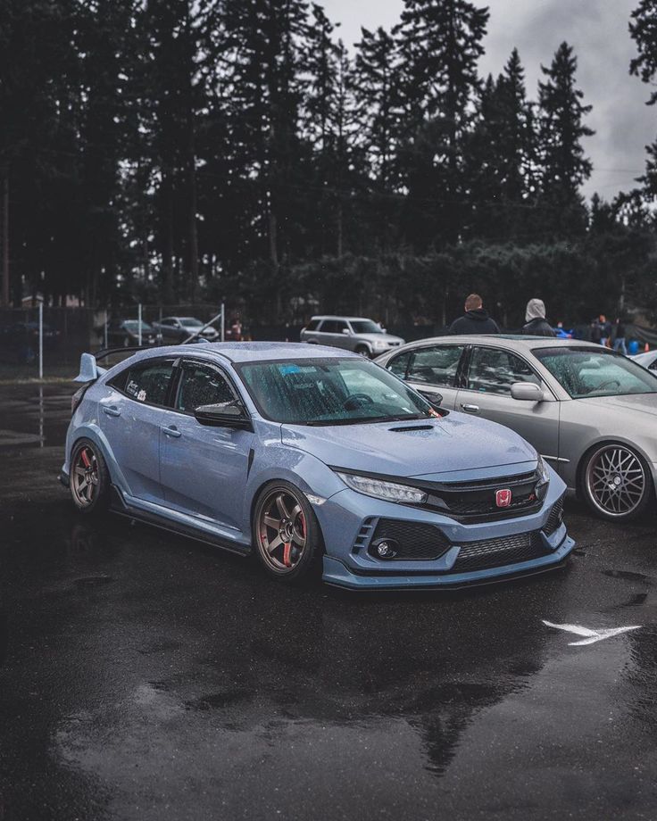 two cars parked in a parking lot with people standing around them and trees behind them