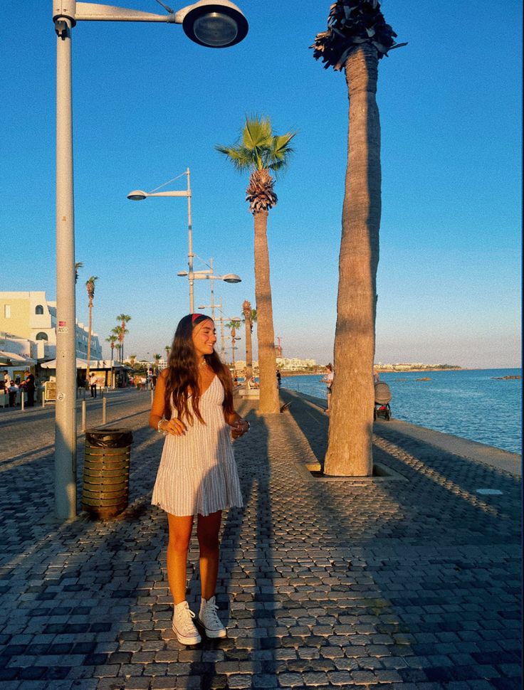 a woman standing on the side of a road next to palm trees and water in front of a street light