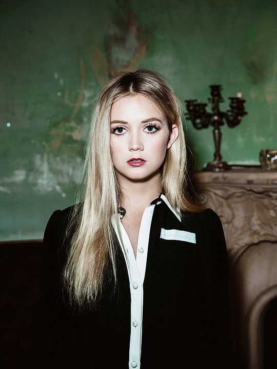 a woman with long blonde hair standing in front of a fireplace and green wallpaper