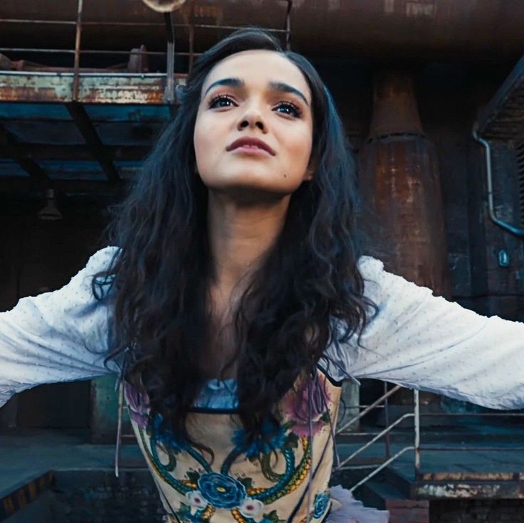 a woman with her arms outstretched in front of an industrial building and pipes behind her