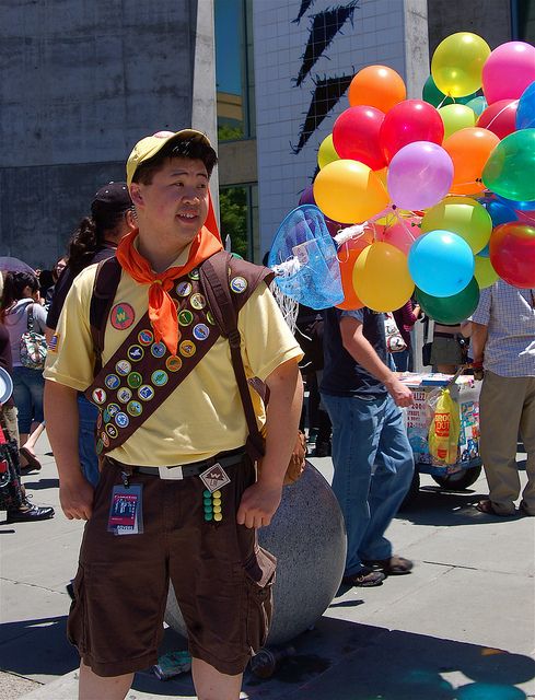 a young man in costume holding balloons and water bottle at an outdoor event with other people