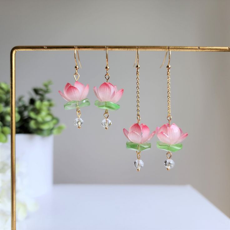 three pink flowers are hanging from gold earwires on a white table next to a potted plant