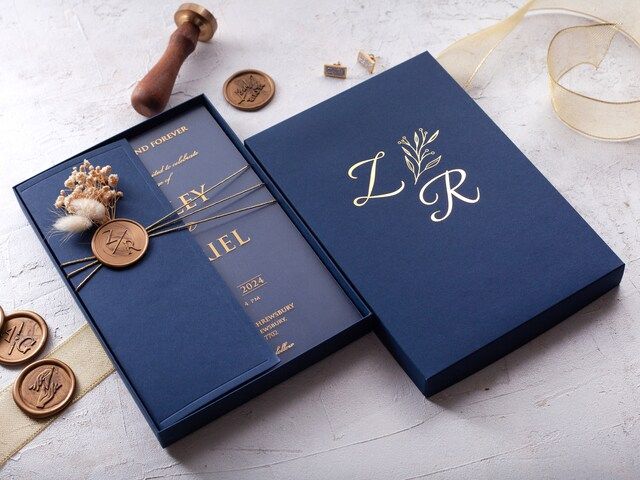 two blue wedding cards with gold initials and ribbons next to some other items on a table