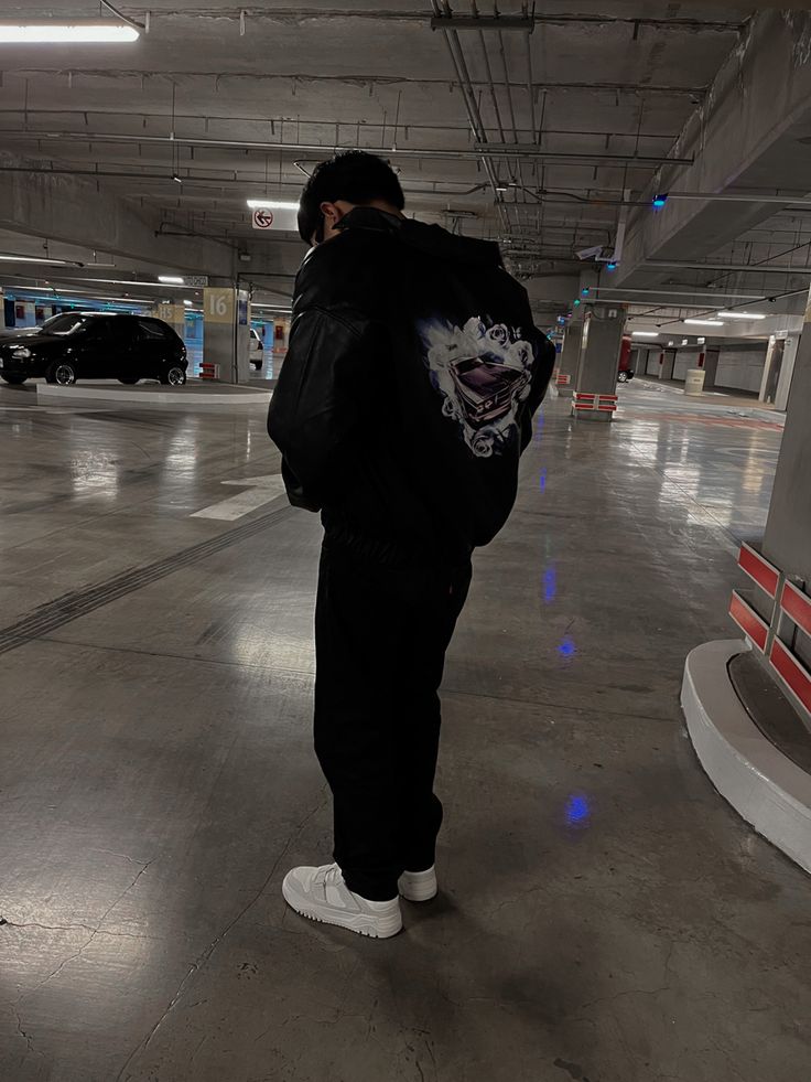 a man standing in an empty parking garage with his back turned to the camera, wearing a black hoodie and white sneakers