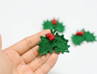 a hand is holding some green crocheted holly leaves with red berries on them