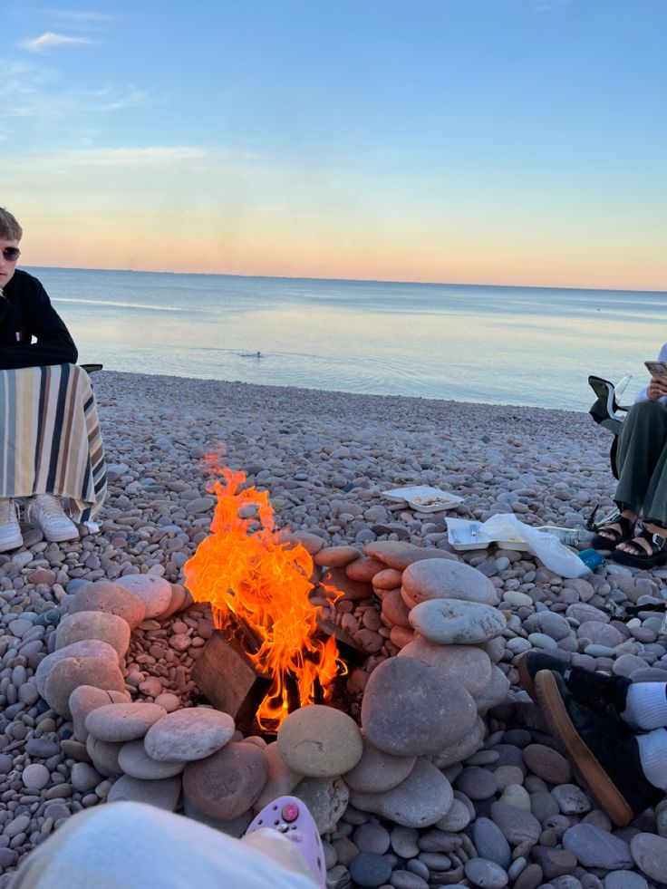 two people sitting around a campfire on the beach