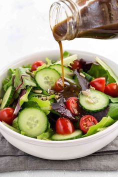 a salad with dressing being poured onto it and topped with cucumbers, tomatoes, lettuce, red onion, and cherry tomatoes in a white bowl