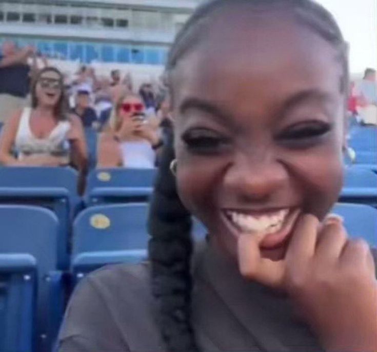 a woman with braids smiling in front of blue seats at a baseball game,