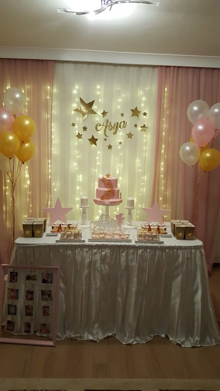 a table topped with a pink cake and lots of gold balloons next to a curtain