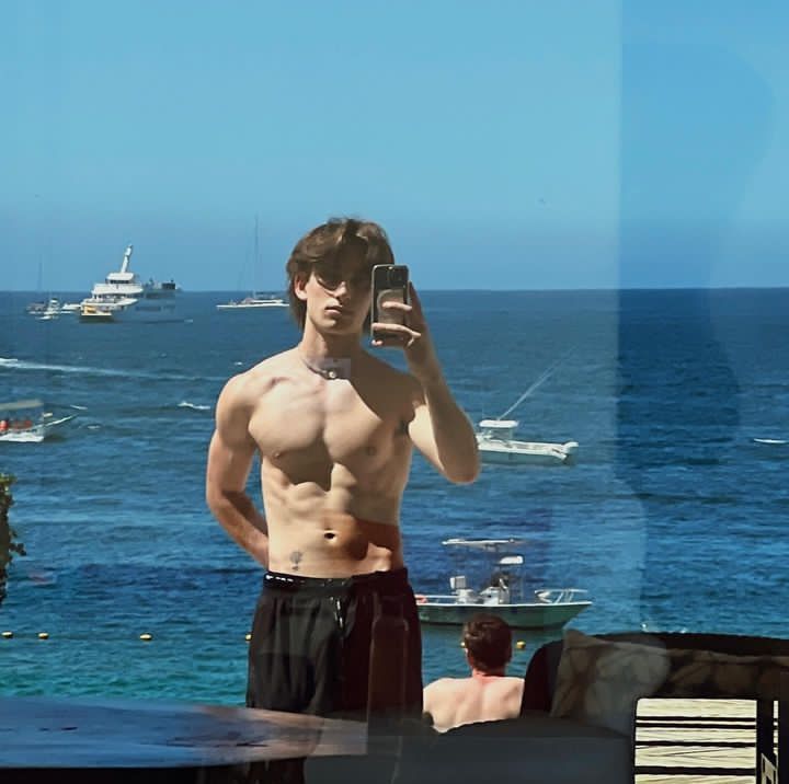 a shirtless man taking a selfie in front of the ocean with his cell phone