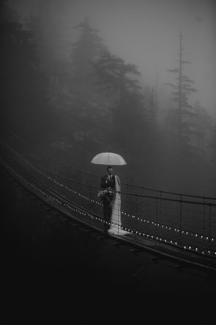 a bride and groom standing under an umbrella on a bridge in the foggy forest