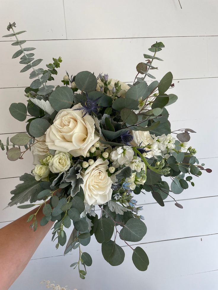 a bouquet of white roses and greenery is held up by someone's hand