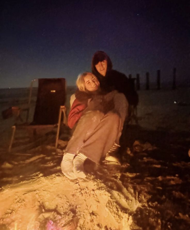 three people sitting around a campfire on the beach at night with their arms around each other