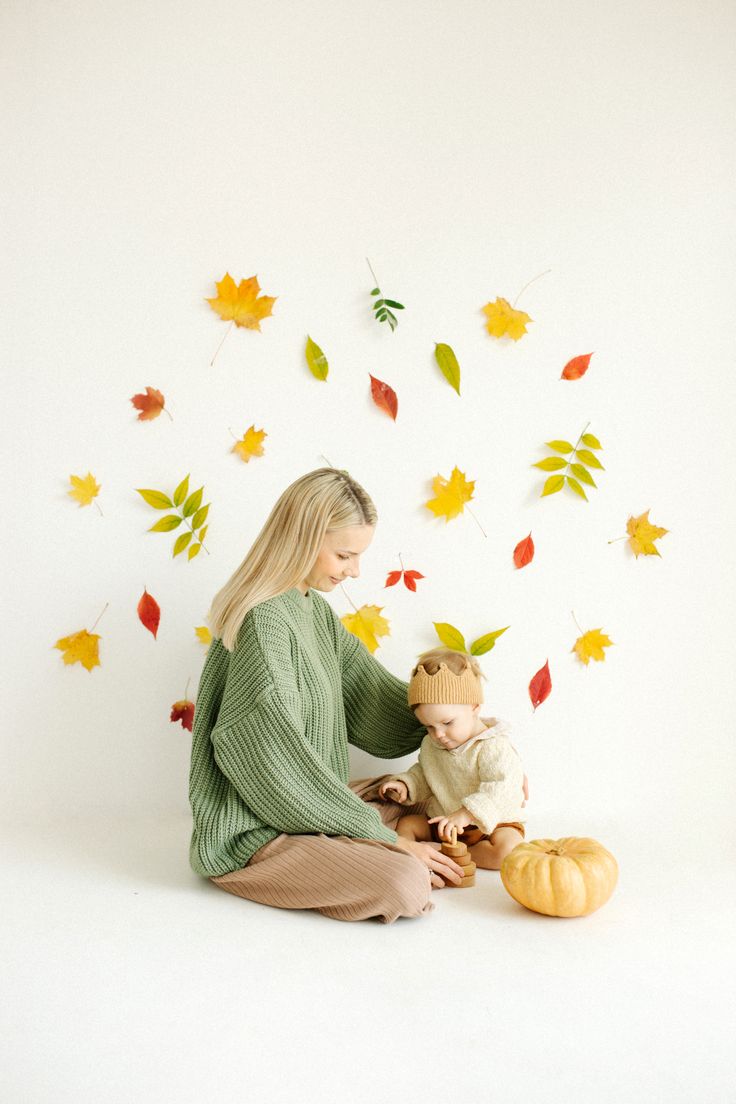 a woman sitting on the floor with her child in front of leaves and pumpkins