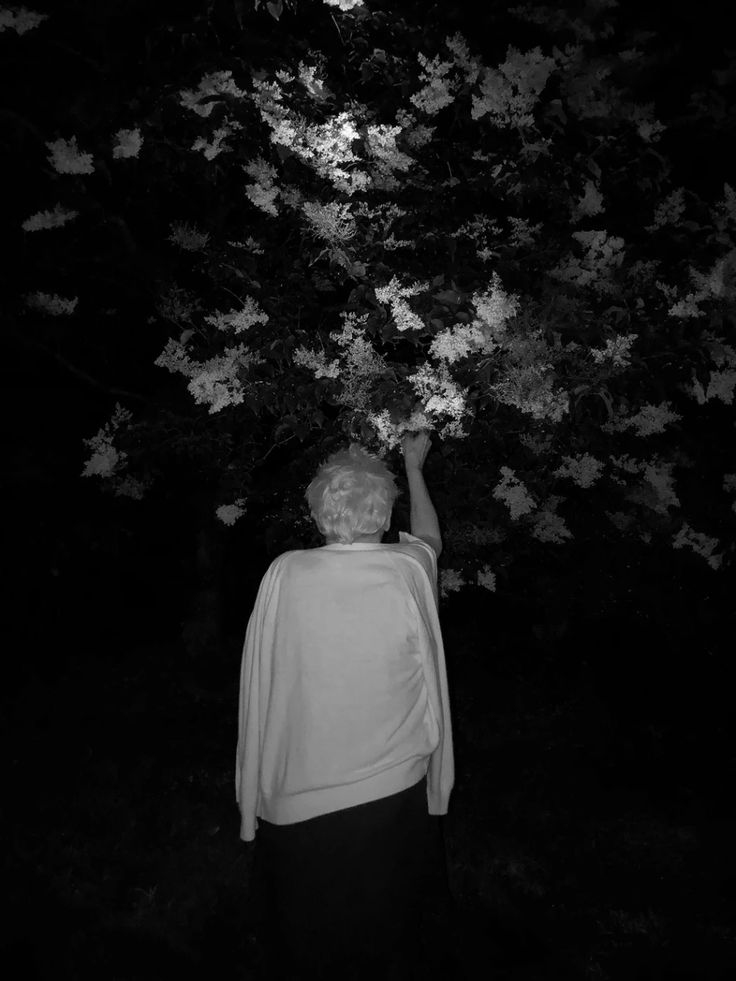 an old woman reaching up to a tree with leaves on it in the night time