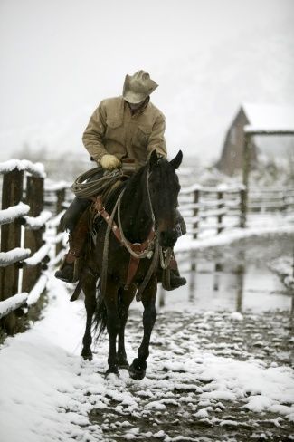 a man riding on the back of a brown horse across snow covered ground next to a wooden fence