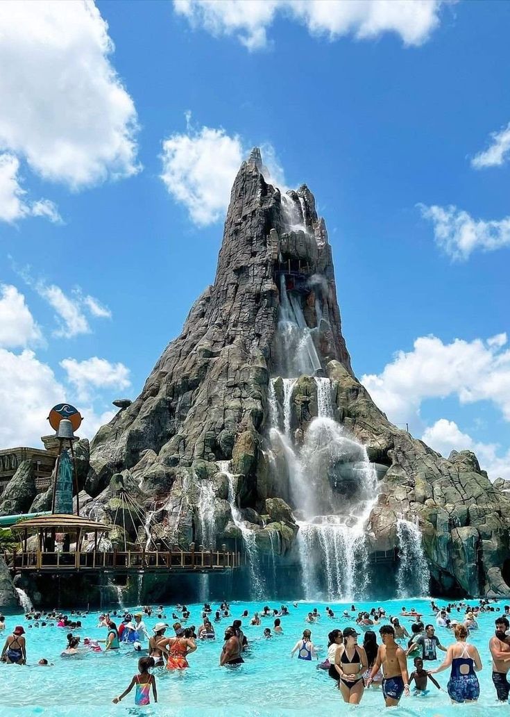 many people are swimming in the water near a large rock formation with a waterfall coming out of it