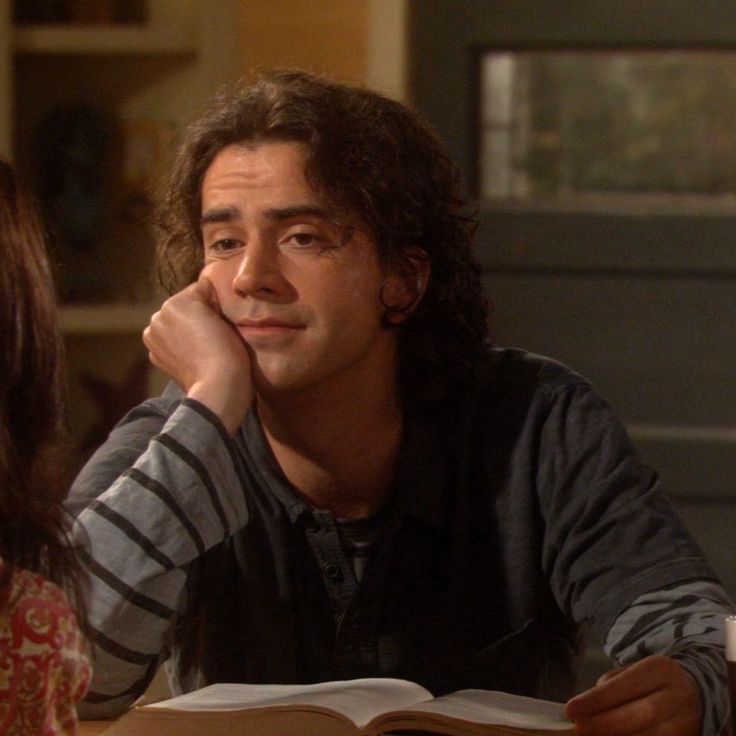 a man sitting at a table with an open book in front of him and another woman looking on