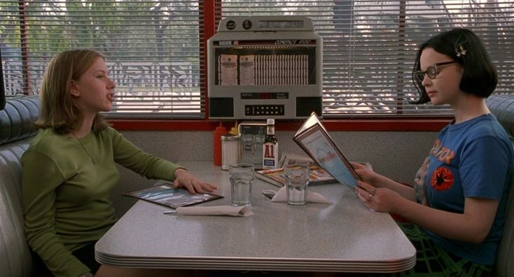 two women sitting at a table talking to each other in front of a window with blinds