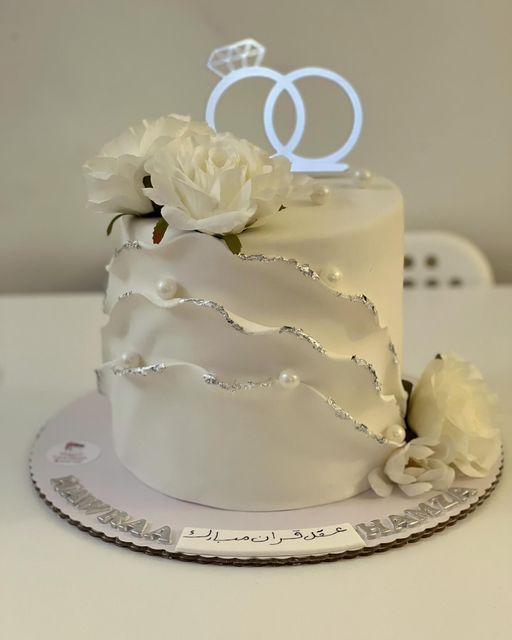 a wedding cake with white flowers and two rings on top, sitting on a table