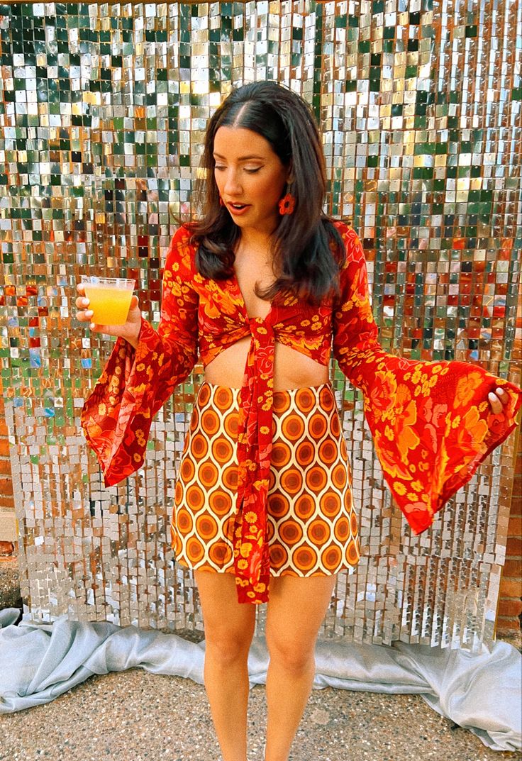 a woman standing in front of a mosaic wall holding a drink and wearing an orange dress