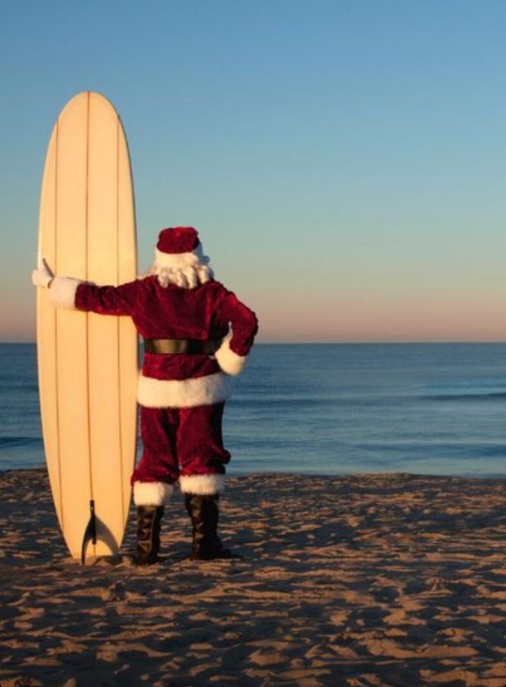 a man dressed as santa claus holding a surfboard on the beach at christmas time