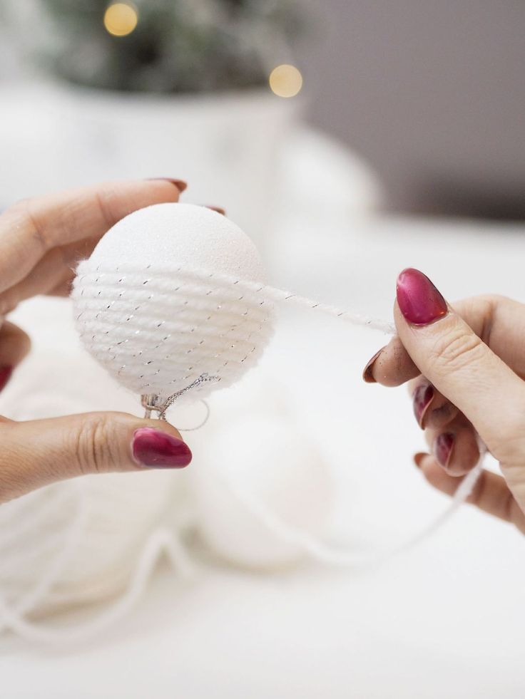 two hands holding a white ball ornament with string attached to it and another hand reaching for one