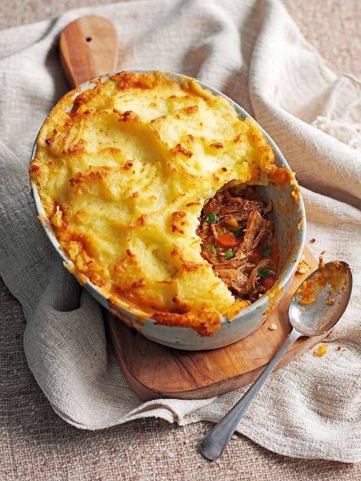 a casserole dish with meat in it on a wooden cutting board next to a spoon