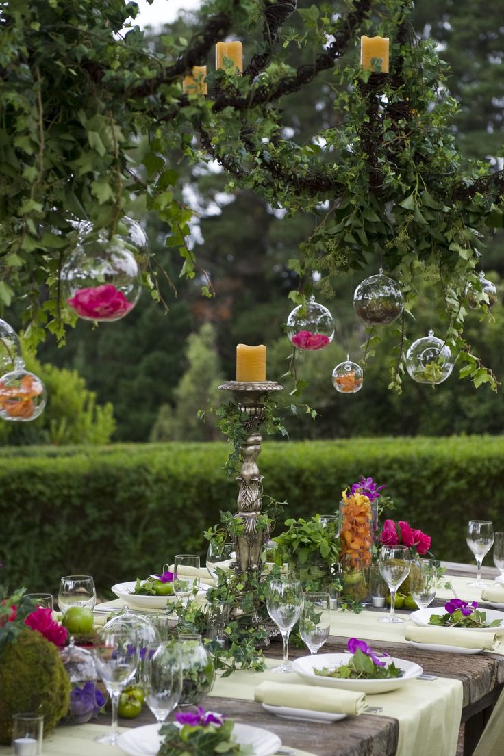 an outdoor dining table with candles, plates and flowers in the center is surrounded by greenery