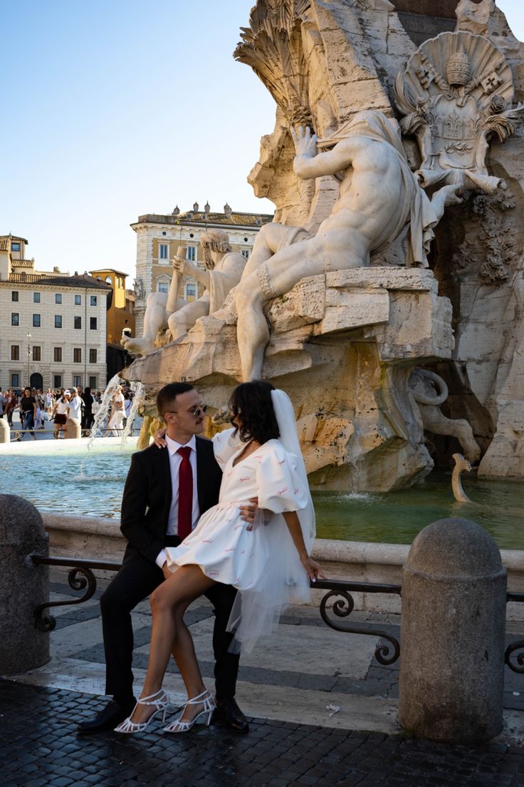 a bride and groom sitting on a bench in front of a fountain with statues behind them