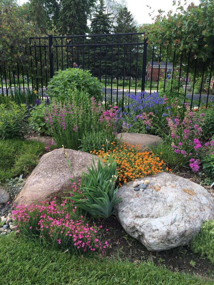 a garden with rocks, flowers and plants in the grass near a black iron fence