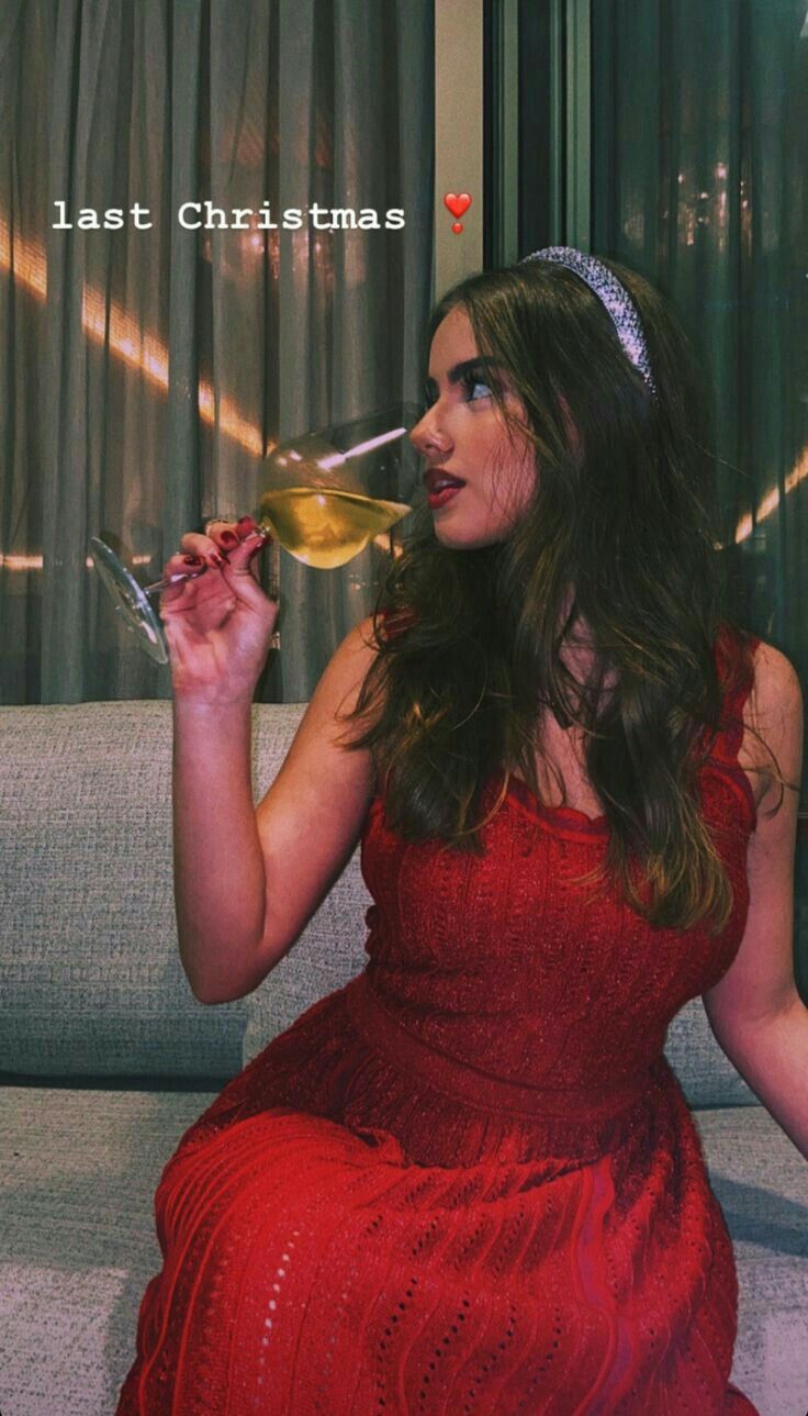 a woman in a red dress sitting on a couch drinking from a wine glass while wearing a tiara