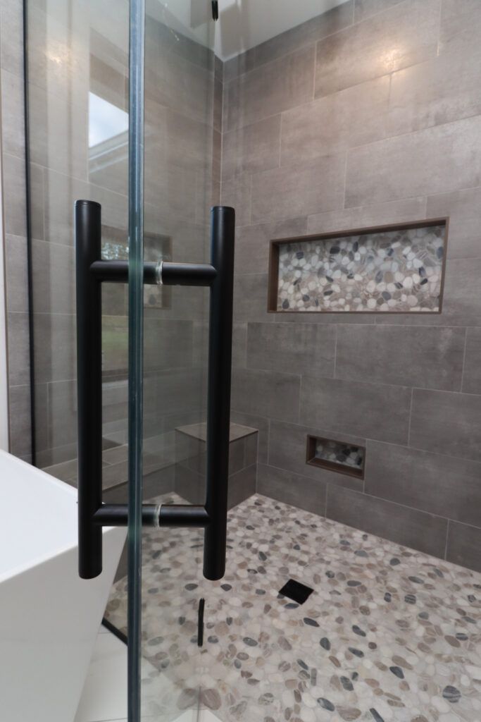 Gray tile shower with light pebble flooring. Glass shower door with black accents. Shower Tile Remodel Ideas, Bathroom Shower Tiles Gray, Gray Tile Master Bath, Gray Tiled Bathrooms Ideas, Grey Tile Shower Ideas Master Bath, Large Gray Tile Shower Ideas, Master Bath Grey Floor, Master Shower Dark Tile, Tile Shower Color Schemes