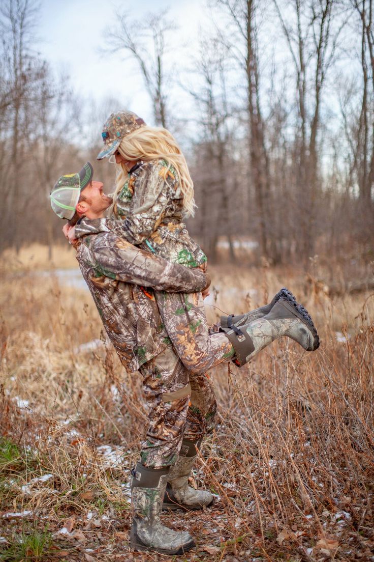 a woman holding a baby in her arms while standing on the ground with trees behind her