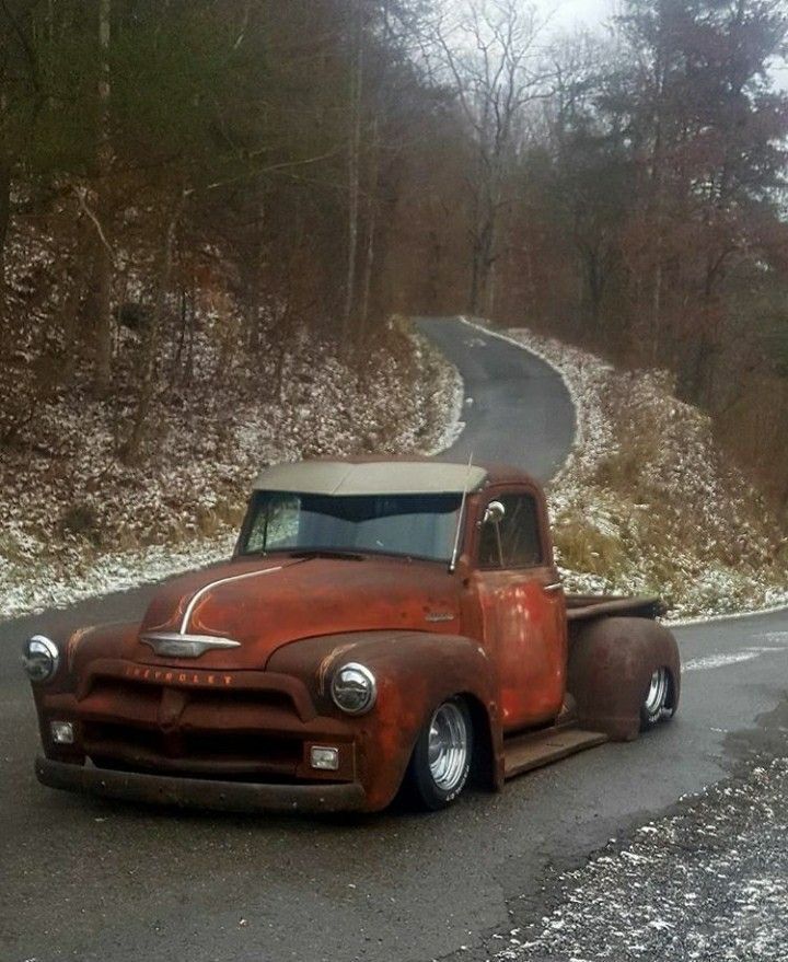 an old rusted pickup truck driving down a road in the woods with snow on the ground