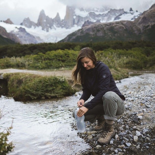 a woman crouches down to collect water from a stream with mountains in the background