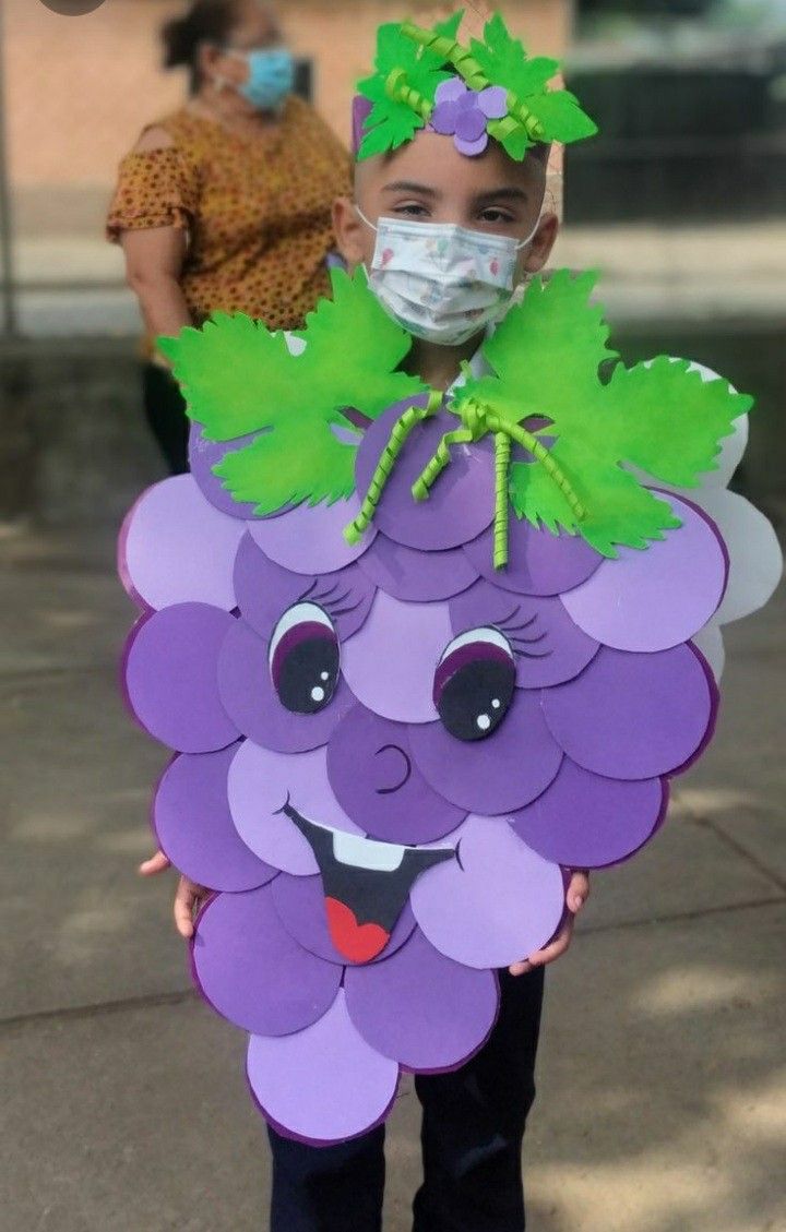 a young boy wearing a face mask and holding a purple grape shaped cutout with green leaves on it